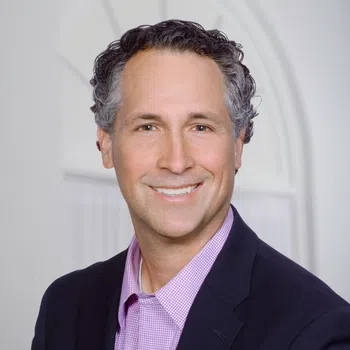 Smiling middle-aged man with curly hair wearing a dark blazer and pink checkered shirt against a light background.