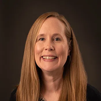 Smiling woman with long blonde hair wearing a black top and silver necklace against a dark background