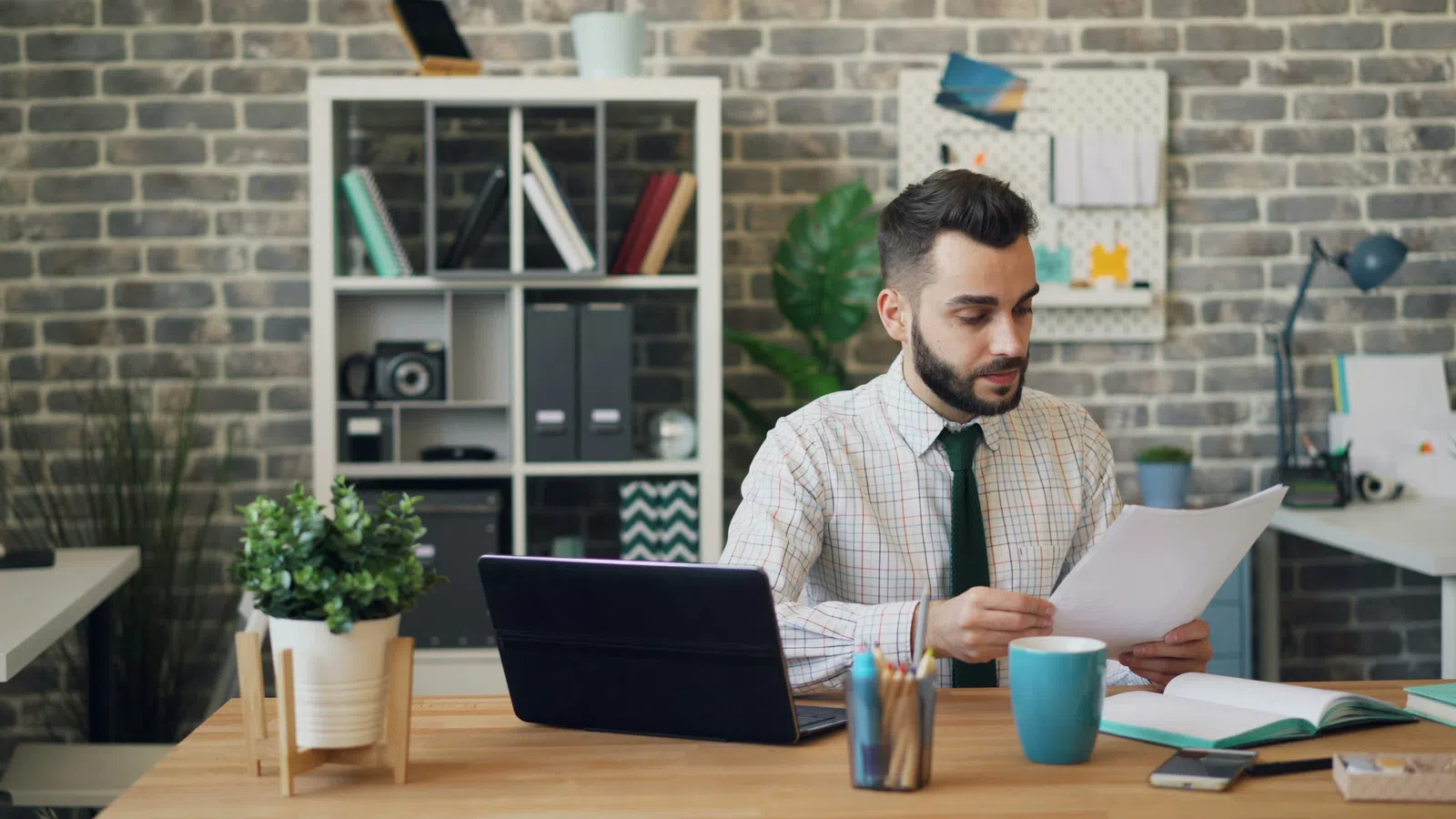 Man in office reviewing documents at desk with laptop, coffee mug, and plants in modern workspace.